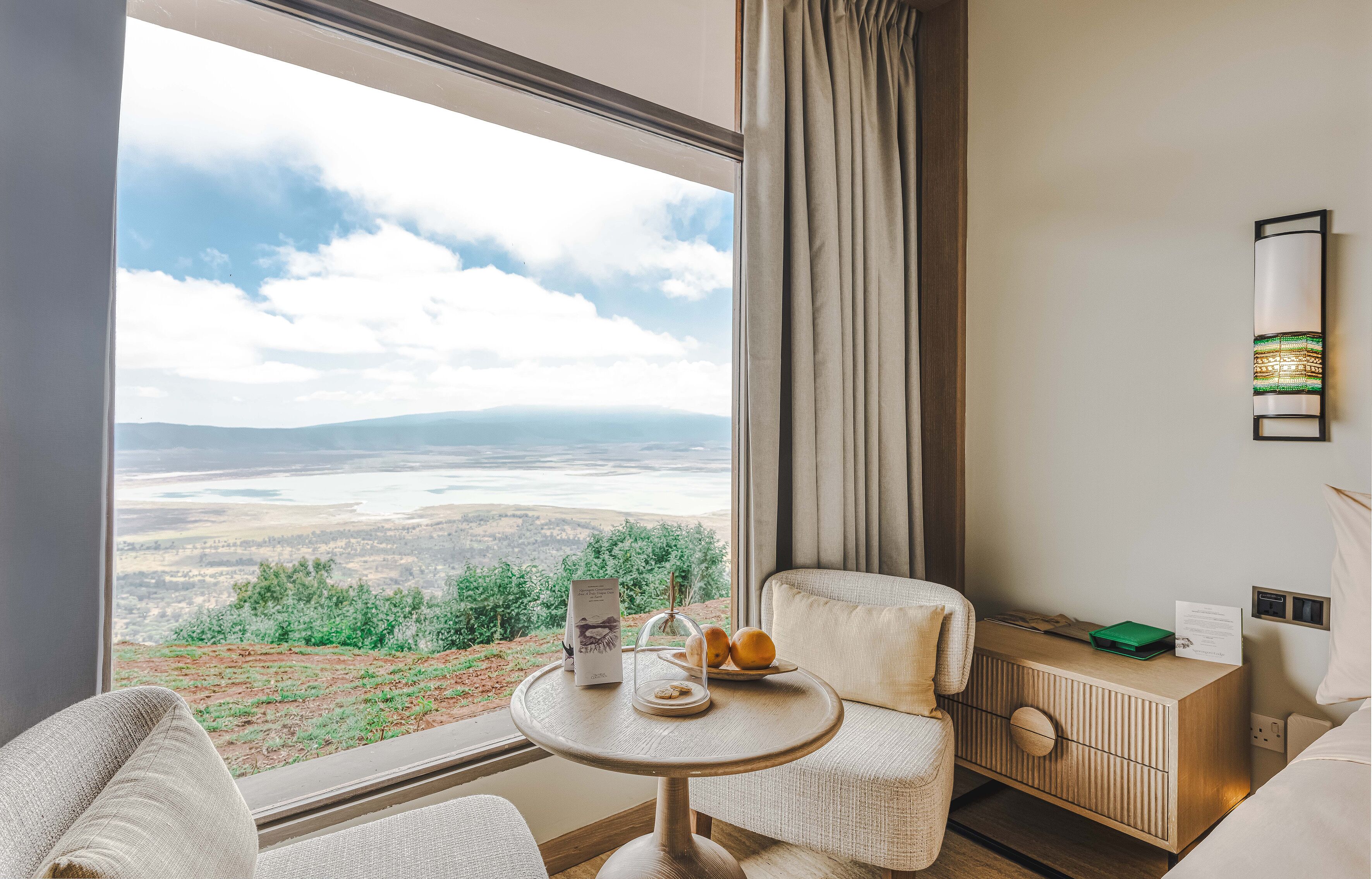 Ein Zimmer mit einem großen Fenster und Blick auf ein Tal und Berge. Ein kleiner runder Tisch mit einer Speisekarte und einer Fruchtschale.