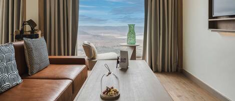 A modern hotel room with a leather sofa, a small table with a decorative item, a green vase, and a view of the landscape through large windows.