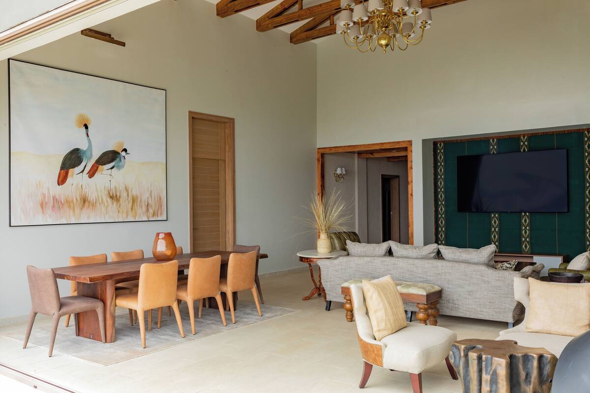 A spacious living room with a wooden dining table, comfortable seating, a large painting, and a flat-screen TV mounted on a green wall.