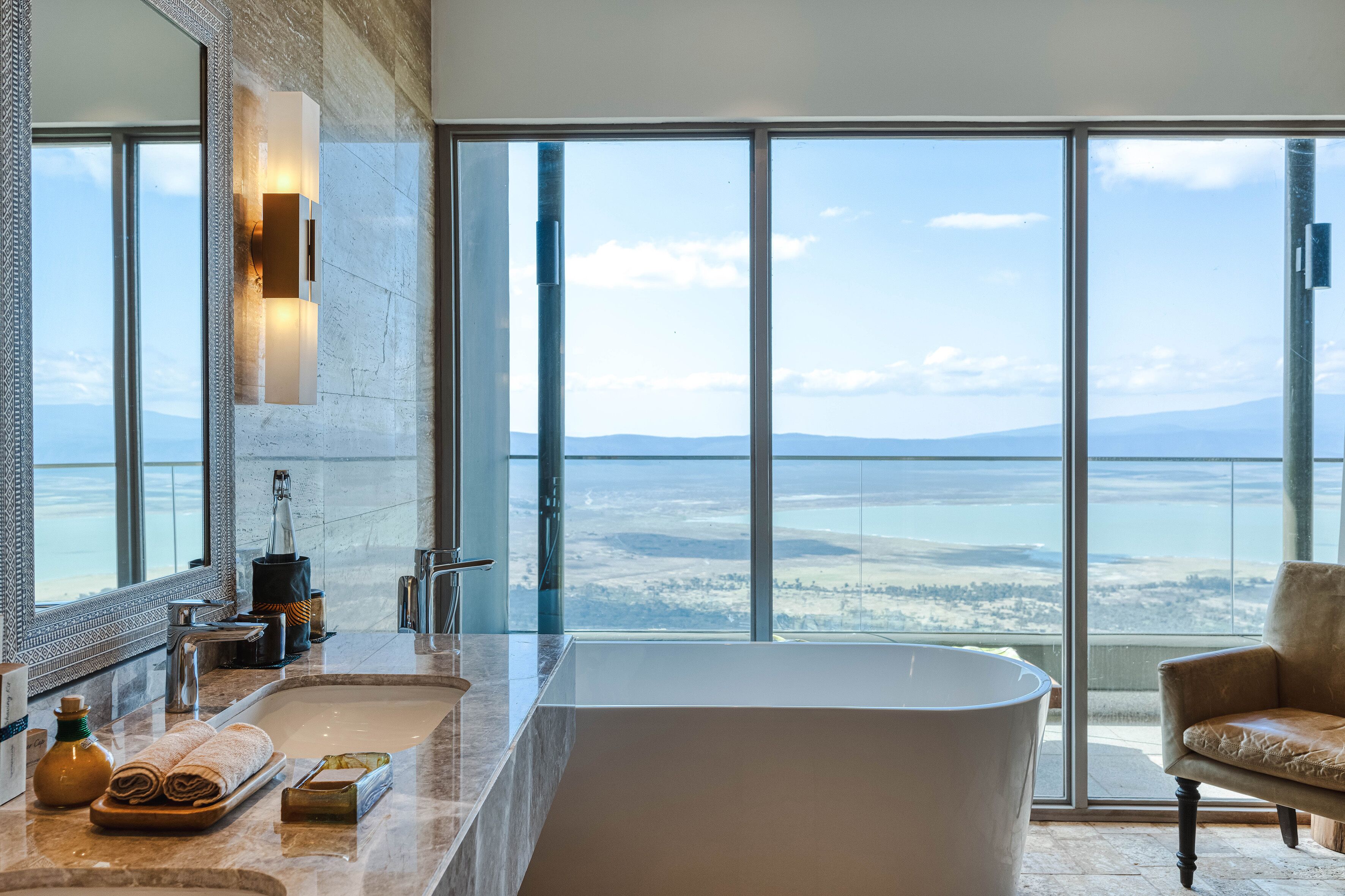Un baño moderno con una bañera grande, un espejo amplio y vistas a un paisaje lejano a través de ventanas grandes.