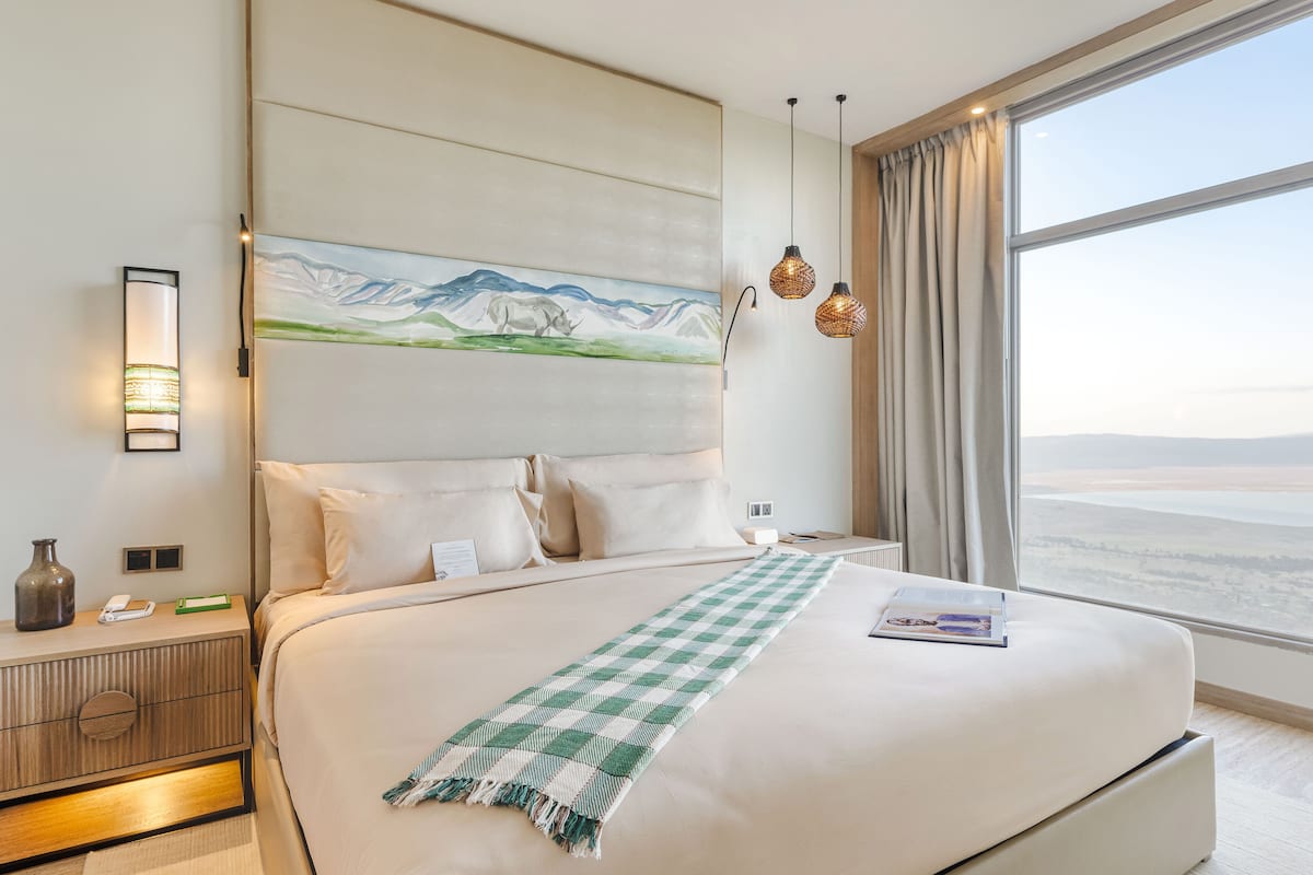 A hotel room with a large bed, a painting on the wall, a nightstand, and a view of the landscape through the window.