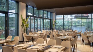 Un comedor amplio con varias mesas y sillas, ventanas grandes que ofrecen vistas al exterior y plantas en macetas que aportan verdor.