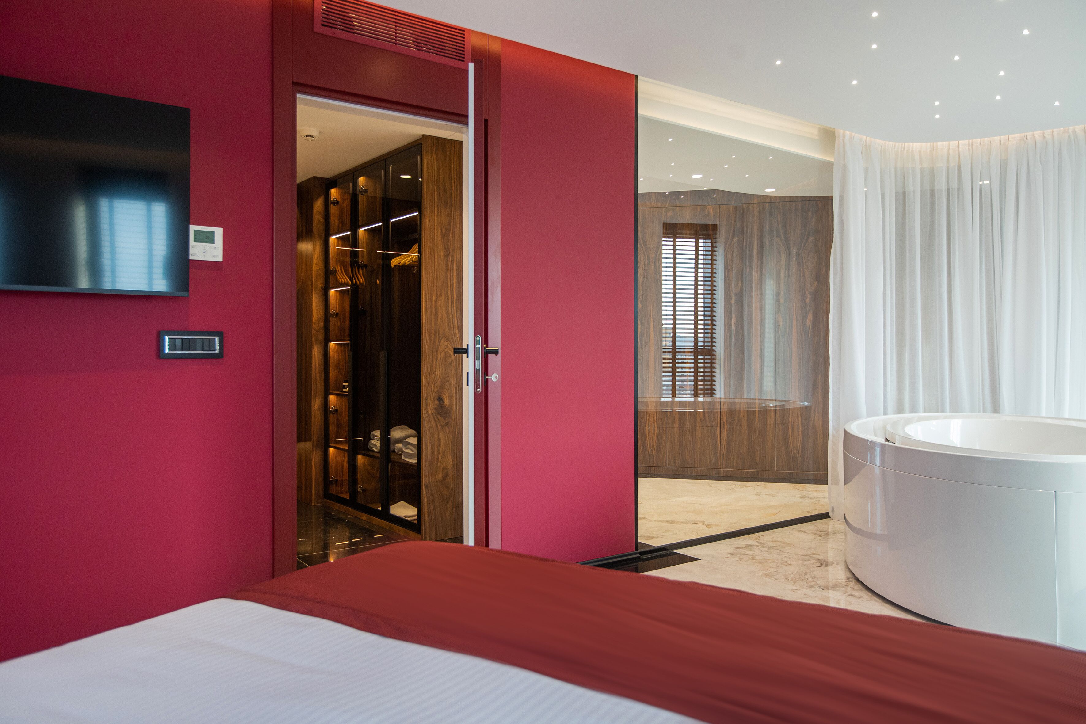 A modern hotel room with a large bed, a walk-in closet, a bathroom with a bathtub, and a TV mounted on the wall.