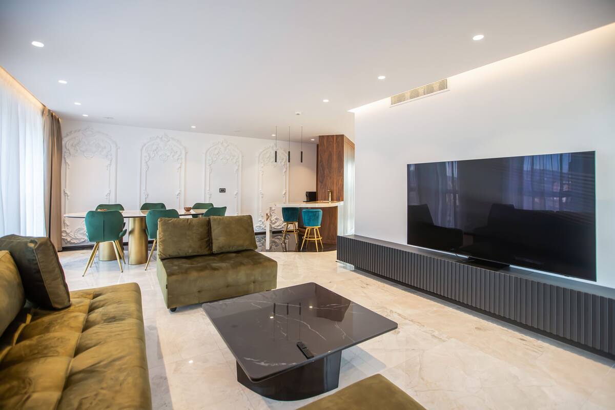 A modern living room with a large flat-screen TV, plush sofas, a marble coffee table, and a dining area with green chairs.