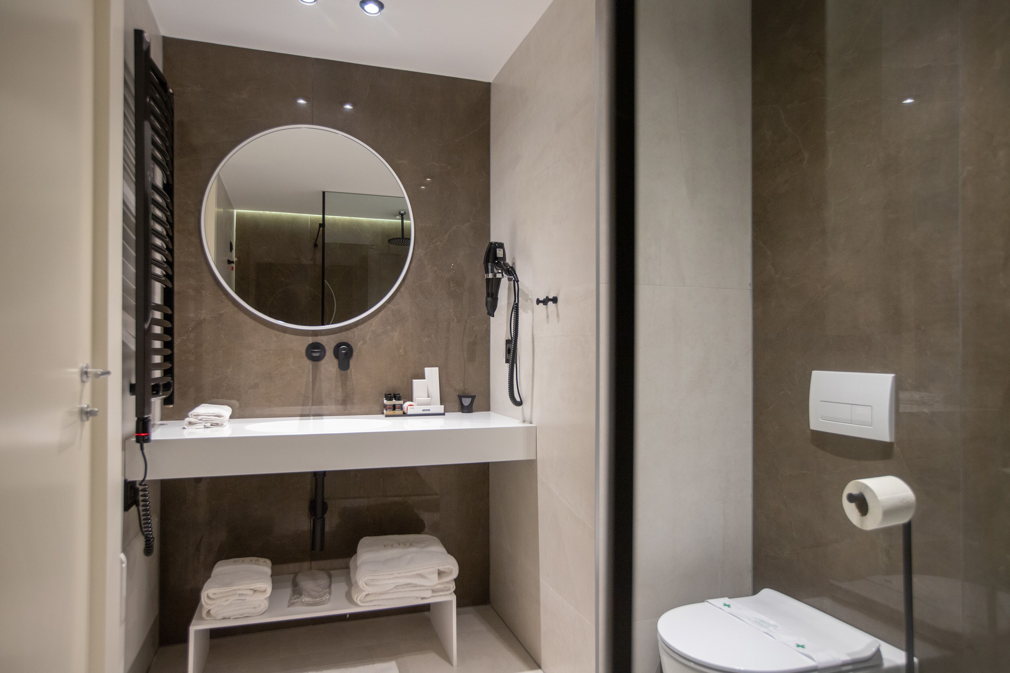 A modern bathroom with a round mirror, a white sink, a handheld showerhead, and a toilet with a paper towel dispenser.