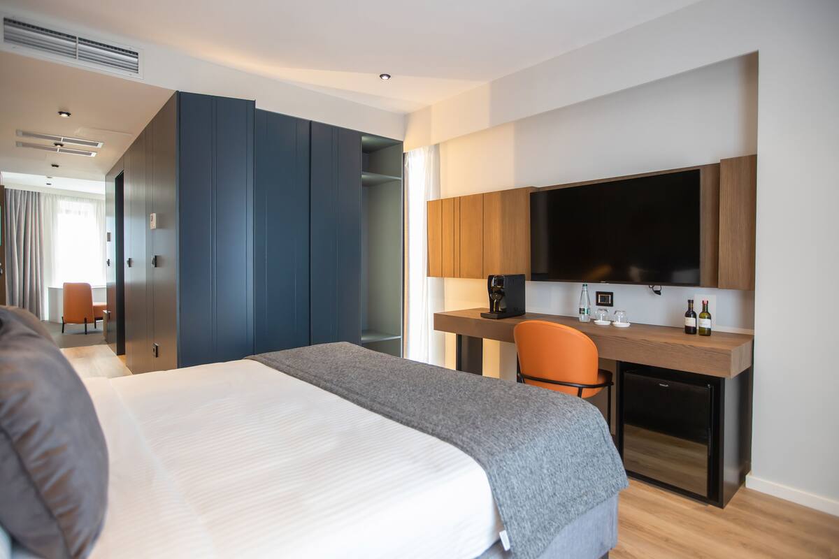 A modern hotel room with a large bed, a desk with a flat-screen TV, and a compact kitchenette.