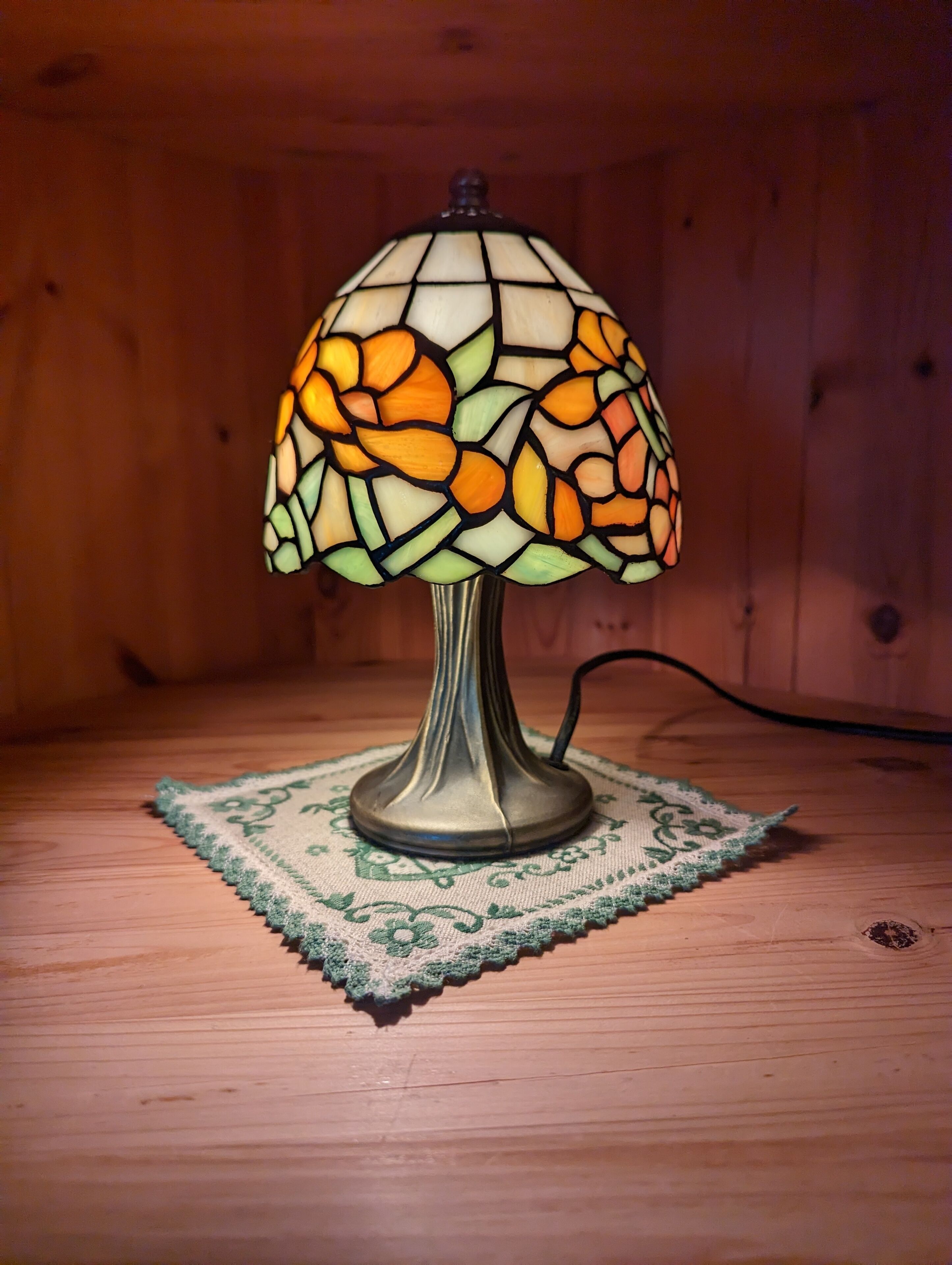 Una lámpara de mesa con una pantalla de vidrio decorada con motivos florales sobre un tablero de madera.