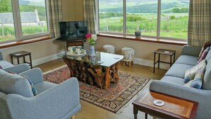 A cozy living room with a wooden coffee table, grey sofas, and a television. Large windows offer a view of green fields and hills. - 4 bedroom accommodation in Advie, near Grantown-on-Spey (Advie, near Grantown-on-Spey)