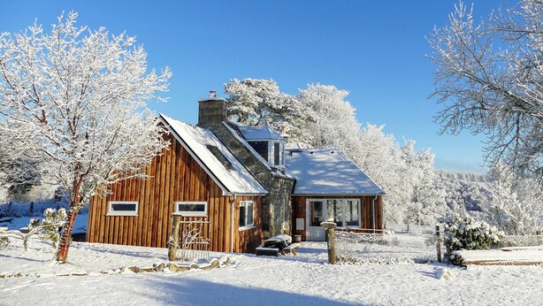 A wooden cabin with a stone chimney, surrounded by snow-covered trees and a clear blue sky. - 4 bedroom accommodation in Advie, near Grantown-on-Spey (Advie, near Grantown-on-Spey)