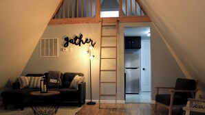 A cozy loft bedroom with a sloped ceiling, a built-in ladder, a kitchenette, and a large window. - A-Frame Mountain Retreat (Gerrardstown)
