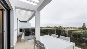 Ett modernt utomhusområde för måltider med ett vitt bord och stolar, ett glasräcke och utsikt över ett bostadsområde.