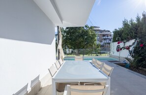 Une terrasse avec une table et des chaises, une piscine et un bâtiment à la façade blanche, avec de la verdure en arrière-plan.
