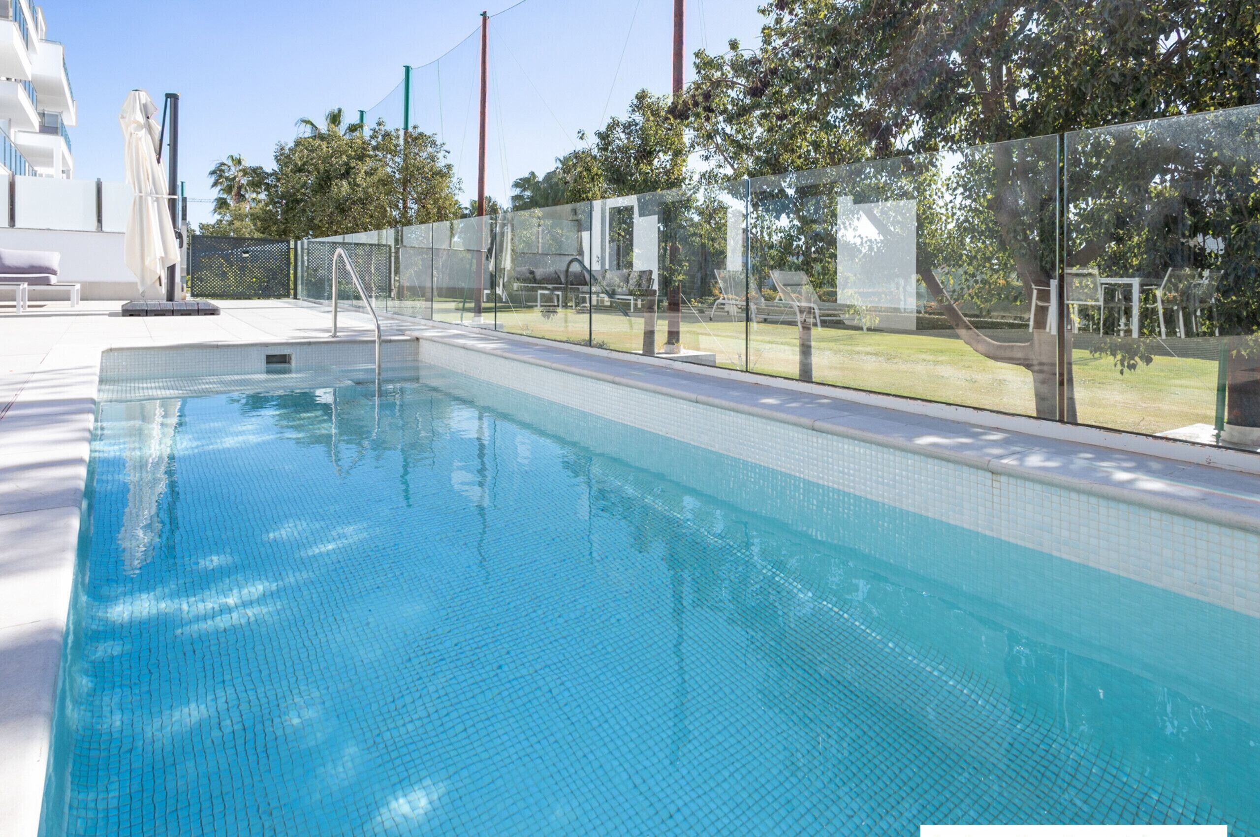 A swimming pool with clear blue water, surrounded by a fence and lounge chairs.