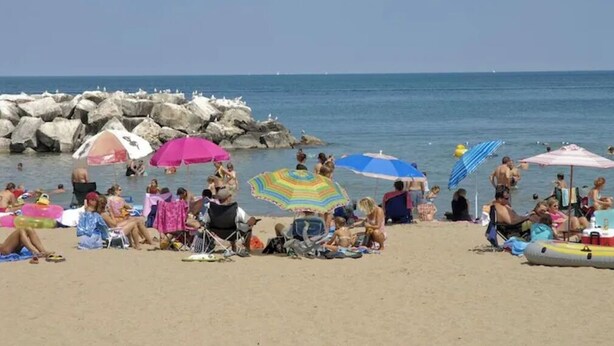 Ein überfüllter Strand mit badenden und sonnenden Menschen, umgeben von bunten Sonnenschirmen.