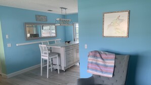 Un comedor con una mesa y sillas blancas, una cocina al fondo y un sillón gris con una manta.