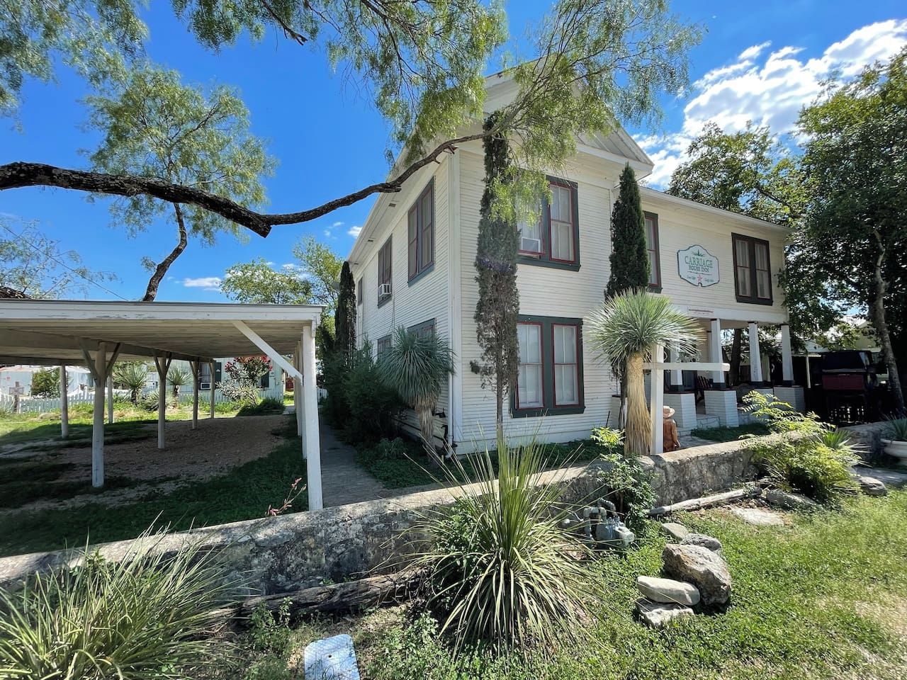 A two-story building with a covered patio area, surrounded by greenery and a stone wall.