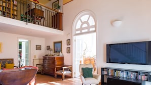 A spacious living room with a high ceiling, a large TV, a bookshelf, a dining table, and a staircase leading to an upper level.