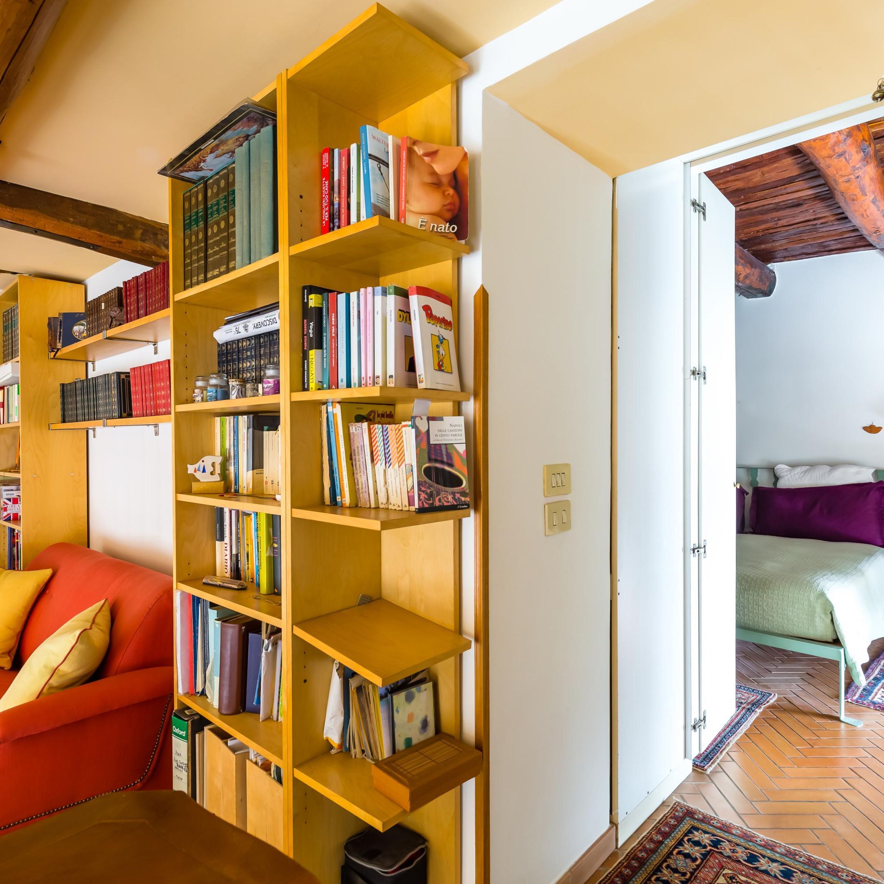 Una habitación acogedora con una estantería de madera llena de libros, un sofá rojo y una cama con un almohá de color morado.
