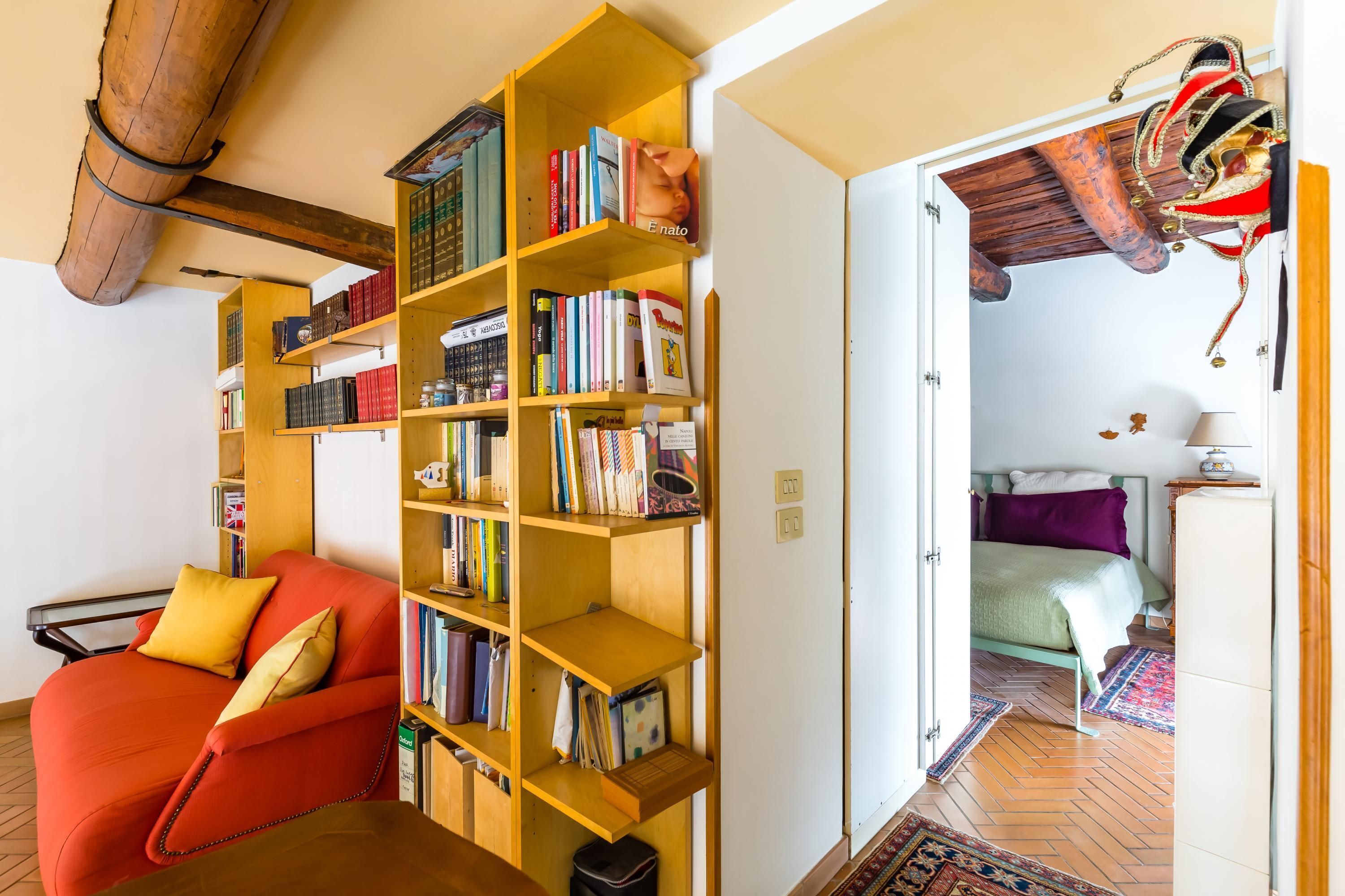 A cozy living room with a red sofa, a wooden bookshelf filled with books, and a view into another room with a bed and a decorative ceiling.