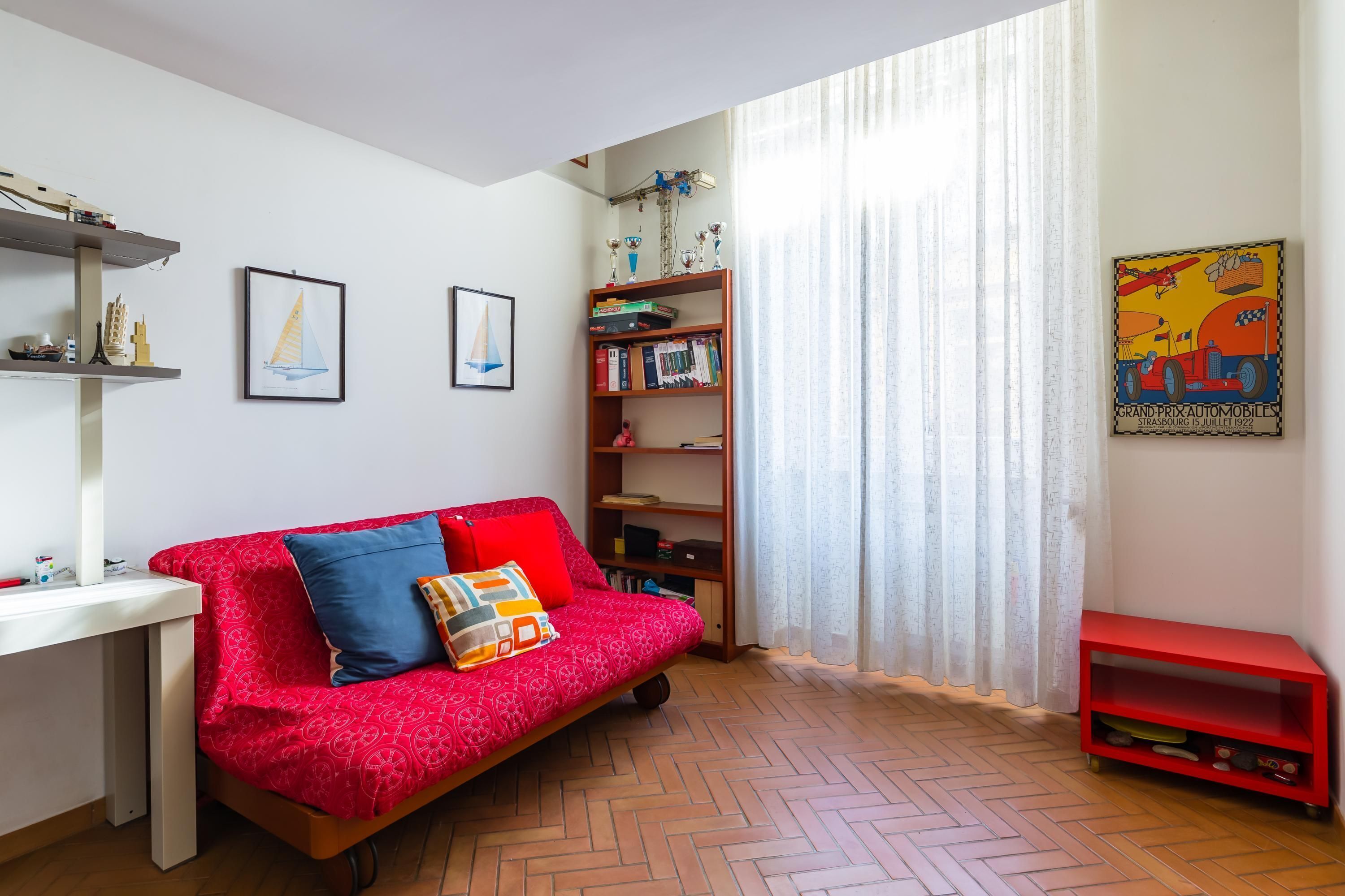 A cozy living room with a red sofa, a white desk, a bookshelf, and a herringbone-patterned floor.