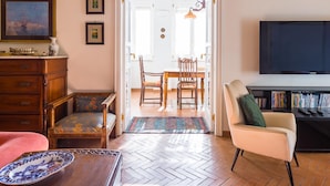 A living room with a wooden floor, a dining area with a table and chairs, a sofa, a television, and a bookshelf.