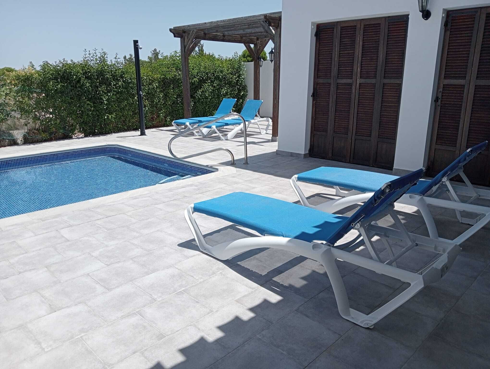 A pool area with blue lounge chairs, a pergola, and a white building with wooden shutters.