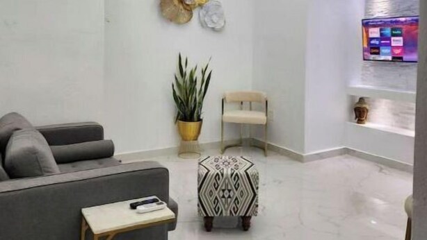 Un área de recepción moderna con un sofá gris, una mesita, un adorno de pared, una planta en maceta y un mostrador de recepción con un monitor de computadora.