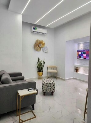 Un espace d’accueil moderne avec un canapé gris, une petite table, un élément décoratif mural, une plante en pot et un comptoir d’accueil doté d’un écran d’ordinateur.