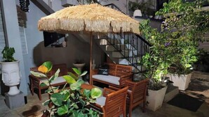 Un área de estar al aire libre con mobiliario de madera y un paraguas de paja, rodeada de plantas en macetas y una escalera.