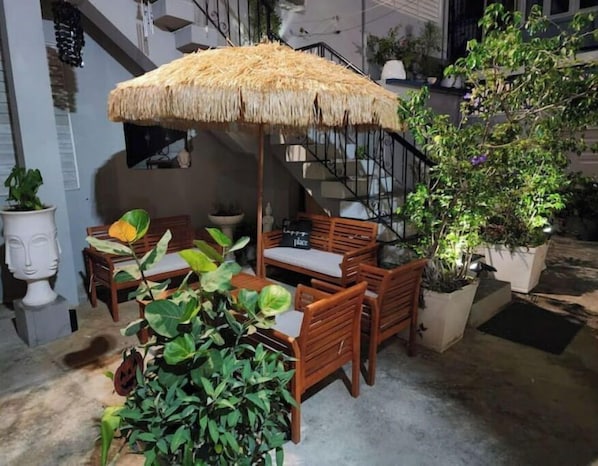 Un espace extérieur aménagé avec du mobilier en bois et un parasol en chaume, entouré de plantes en pot et d’un escalier.