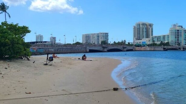 Ein Strand mit einer Brücke im Hintergrund, sandigem Ufer und ruhigem Wasser.