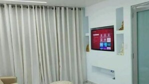 Ein modernes Hotelzimmer mit einem Bett, einem kleinen runden Hocker, einem beige-farbenen Sofa, einem großen Flachbildfernseher an der Wand und einem Vorhang.