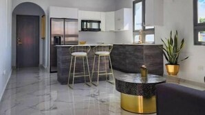 Una cocina moderna con encimera oscura y taburetes de bar, zona de comedor con mesa redonda y sillas, y un ventanal que proporciona luz natural.