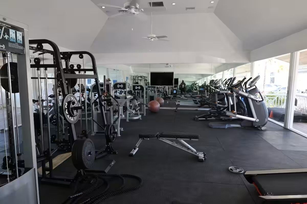 Un gimnasio con diverso equipo de ejercicio, incluyendo cintas de correr y máquinas de pesas, y amplias ventanas que ofrecen vistas al exterior.
