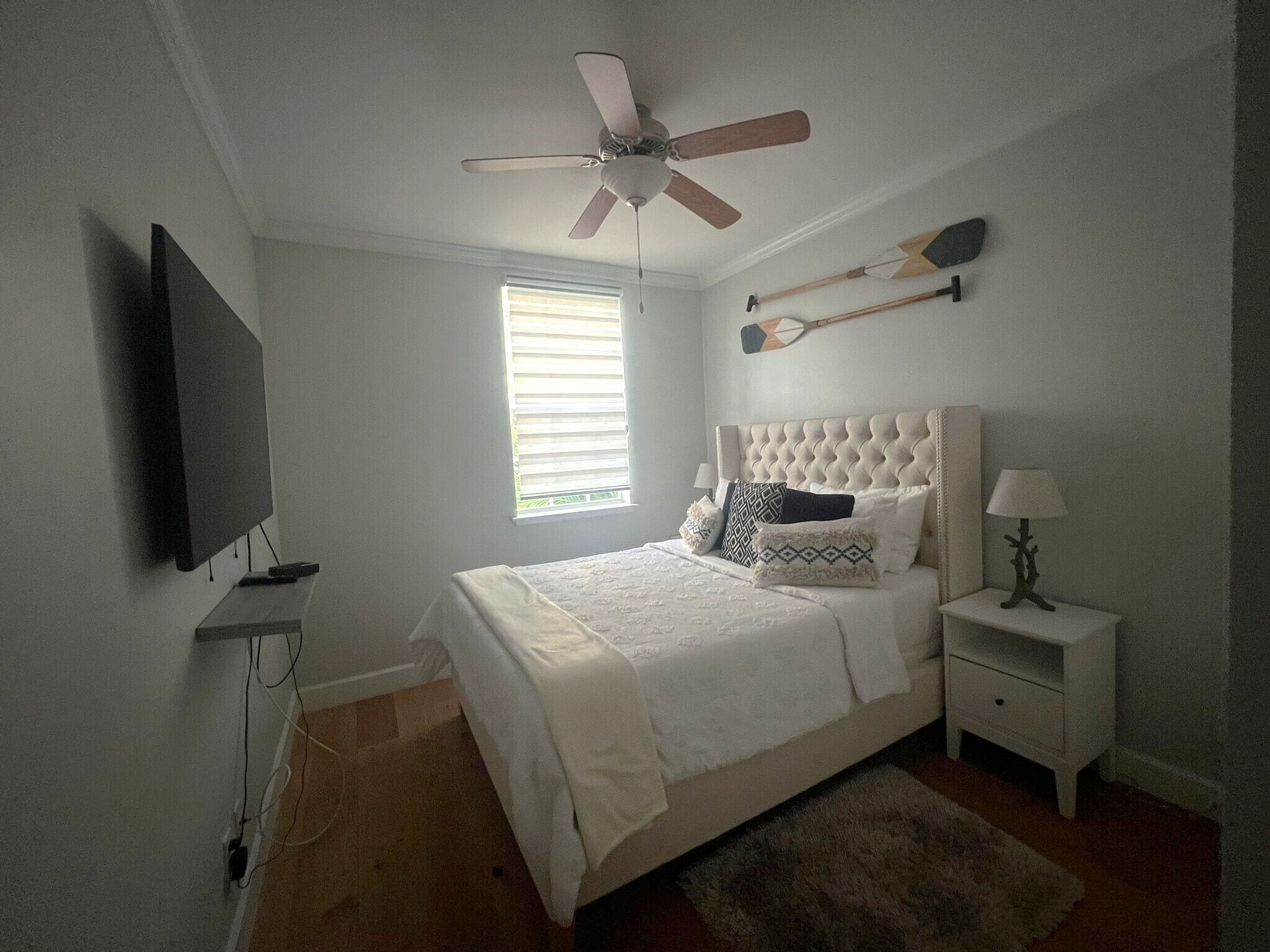Ein Schlafzimmer mit Bett, Deckenventilator, an der Wand montiertem Fernseher, Nachttisch mit Lampe und Fenster mit Jalousien.