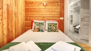 Ein Schlafzimmer mit einem Holzkopfteil, einem Bett mit floralen Kissen und einem grünen Teppich. Eine weiße Tür führt zu einem Kleiderschrank mit Regalen.
