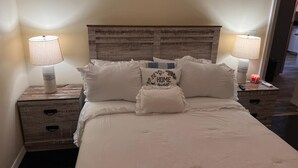 A neatly made bed with white bedding and decorative pillows, flanked by two bedside tables with lamps and a small nightstand with a lamp. - Cute, clean and cozy cottage. 5 minute drive from Junkanoo beach and Bay street. (Nassau)