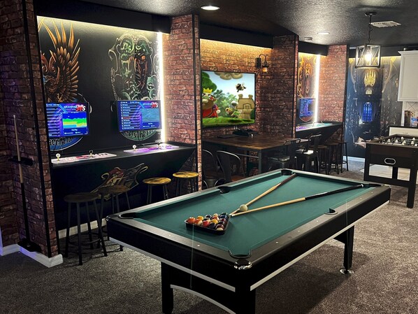 A pool table with balls, a foosball table, and two screens displaying video games.