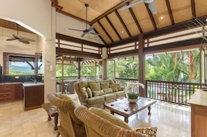 A spacious living area with a balcony, wicker furniture, ceiling fans, and a view of greenery.