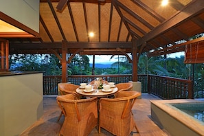 A covered outdoor dining area with wicker chairs and a table set for four, overlooking a tropical landscape.