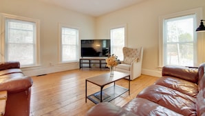 A spacious living room with a brown leather sofa, a white armchair, and a coffee table with a vase of flowers. A flat-screen TV is mounted on the wall, and there are large windows providing natural light. - All-Season Ludlow Apartment: 3 Mi to Okemo Resort! (Ludlow)