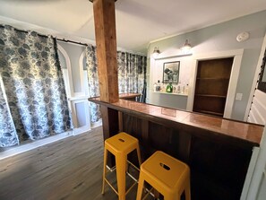 A room with a wooden bar counter, two yellow stools, patterned curtains, a shelf with bottles, and a framed picture on the wall. - Old Florida Charm (Eustis)