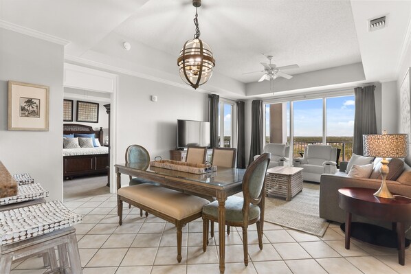 A spacious living area with a dining table, comfortable seating, a large window with a view, and a ceiling fan.