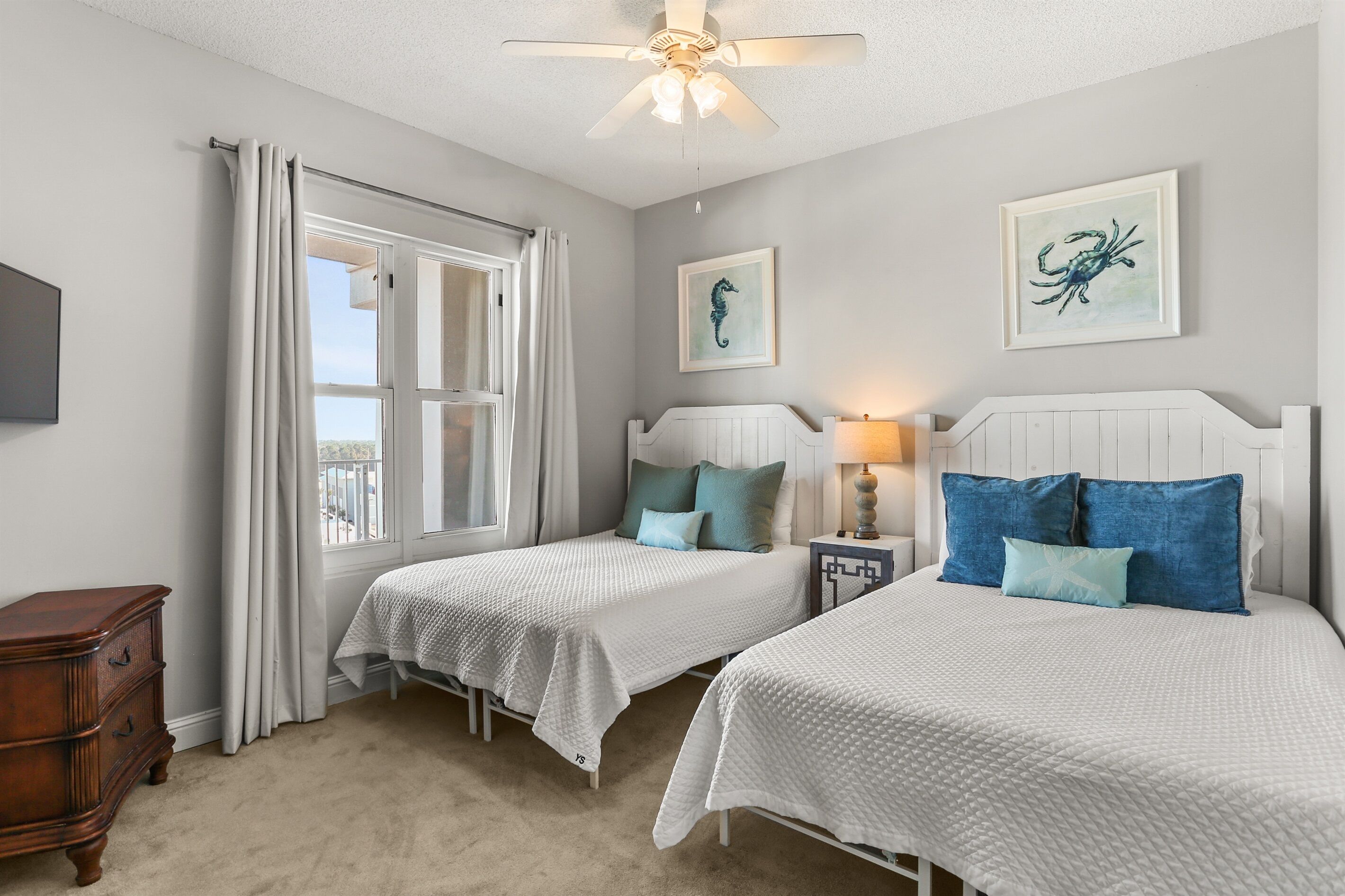 A bedroom with two beds, a ceiling fan, a window with curtains, and artwork on the walls.