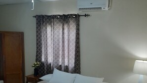 Una habitación de hotel con cama, ventilador de techo, unidad de aire acondicionado, mesita de noche con lámpara y ventana con cortinas estampadas.