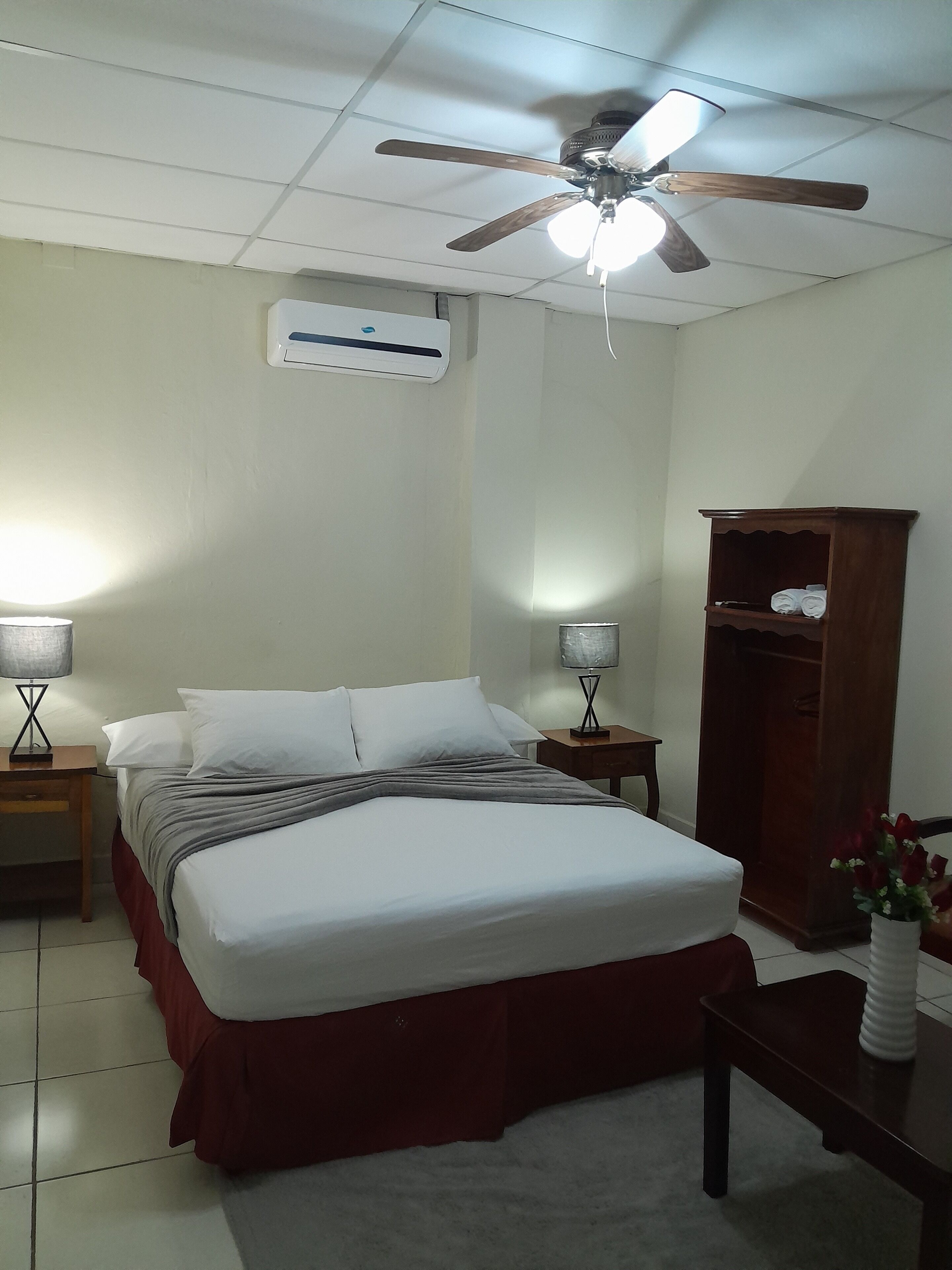 A bedroom with a bed, two bedside tables with lamps, a ceiling fan, an air conditioning unit, and a wooden wardrobe.