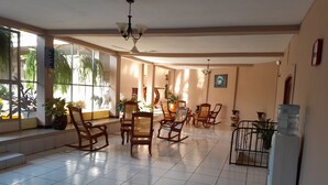 Una habitación amplia con suelo de baldosas, varias sillas mecedoras de madera, una mesa de comedor con sillas y ventanas grandes que dejan entrar luz natural.
