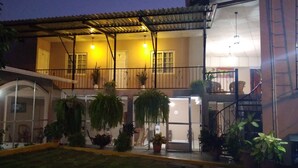 Un edificio de dos pisos con un patio cubierto, varias plantas en macetas y una escalera que lleva a un nivel superior.