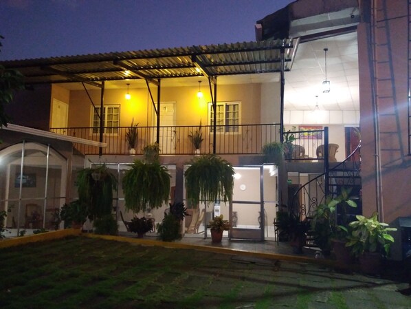 Um edifício de dois andares com um alpendre coberto, várias plantas em vasos e uma escadaria que leva a um andar superior.