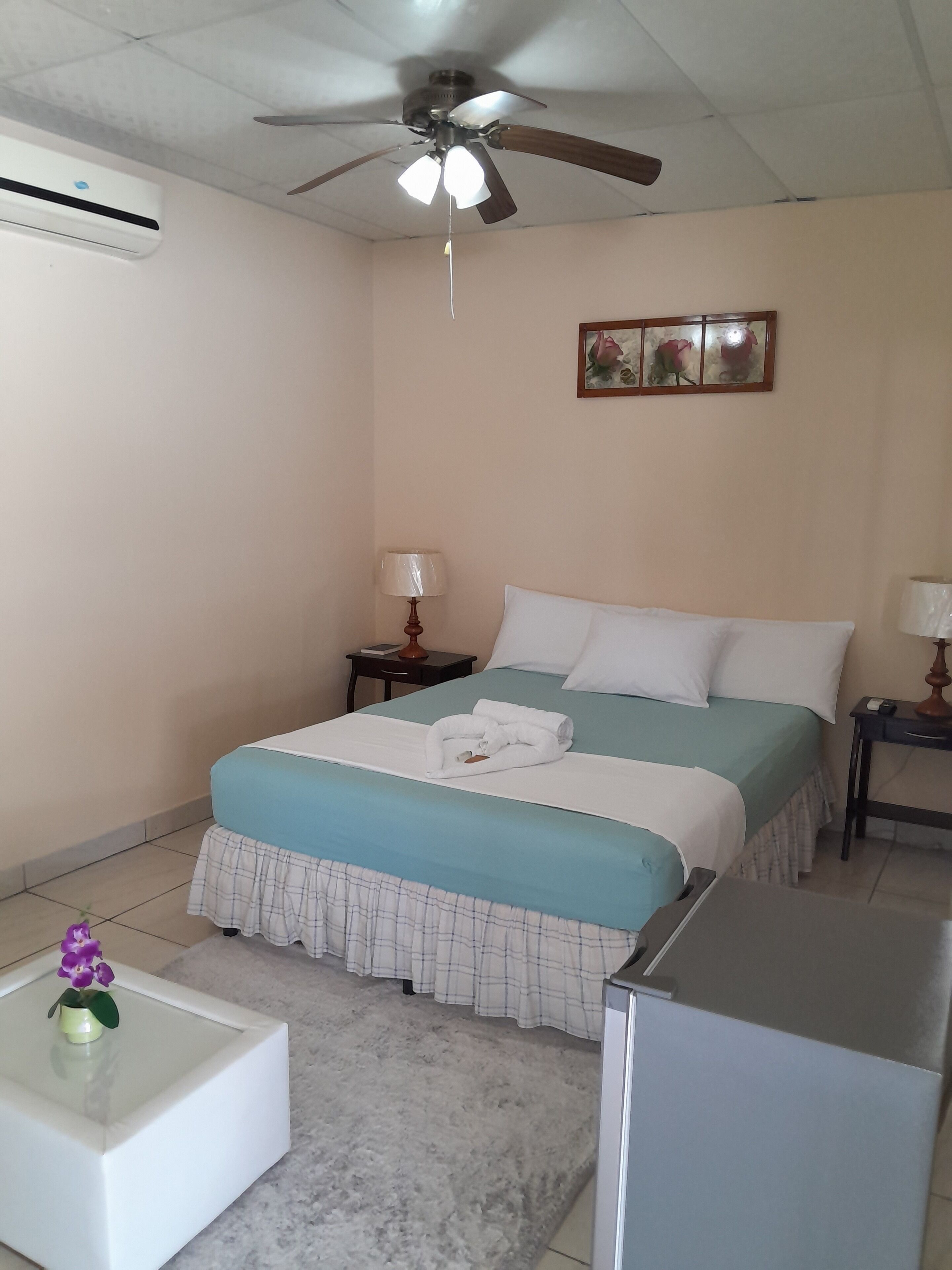 Quarto de hotel com cama, ventoinha no teto, mesa de cabeceira, luminária, pequena mesa com uma flor e um refrigerador.